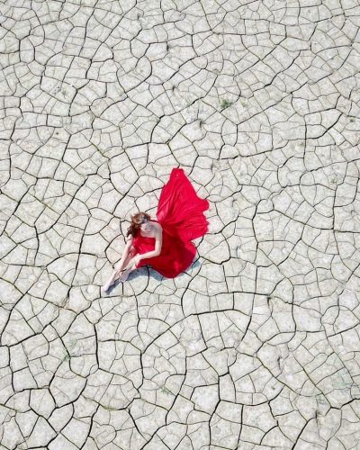 Die Datei ist ein Bild, das eine Frau in einem roten Kleid zeigt, die auf einem ausgetrockneten, rissigen Boden sitzt. Sie ist von oben fotografiert, wobei die Risse im Boden ein auffälliges Muster bilden, das das Bild dominiert. Die Frau sitzt in der Mitte des Bildes und ihr leuchtend rotes Kleid bildet einen starken Kontrast zur erdigen Farbe des Bodens. Ihr Körper ist leicht zur Seite gedreht.